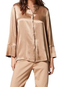 Blusa in seta bronzo con chiusura a bottoni, colletto e maniche ampie. Texture liscia e vestibilità comoda, abbinata a pantaloni beige.