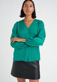 Blusa verde smeraldo con scollatura a V e maniche lunghe a palloncino, abbinata a una minigonna nera in pelle. Tessuto morbido con vestibilità rilassata.