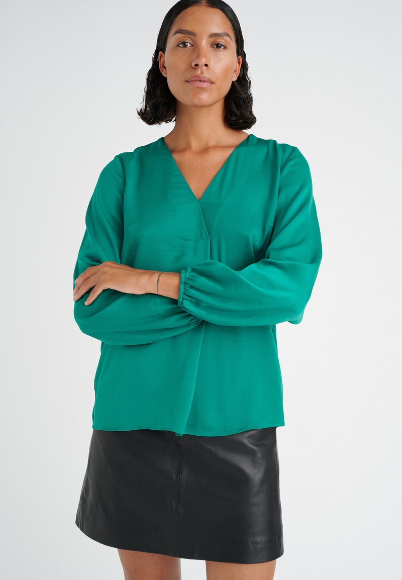 Blusa verde smeraldo con scollatura a V e maniche lunghe a palloncino, abbinata a una minigonna nera in pelle. Tessuto morbido con vestibilità rilassata.