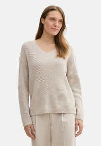 Ljust beige stickad tröja med V-ringning, ribbad textur och chevronmönster. Lätt oversize passform med långa ärmar, kombinerad med krämfärgade byxor.