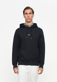 Joven vestido con una sudadera negra de GANT sobre una camisa de cuadros y pantalones blancos, de pie contra un fondo blanco liso.