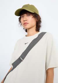 Jaunas žmogus su garbanotais plaukais, dėvintis alyvuogių žalios spalvos kepurę, per didelį beige Nike marškinėlius ir languotą petinę rankinę, prieš baltą foną.