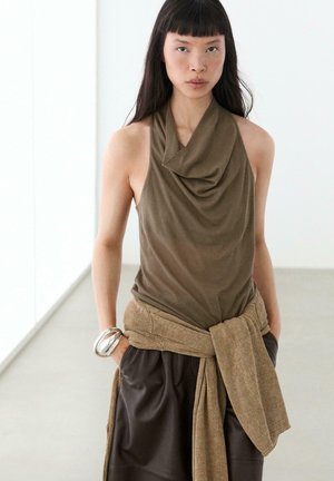Frau mit langen dunklen Haaren trägt ein ärmelloses olivgrünes drapiertes Top, dunkle Hose, beige Strickjacke um die Taille gebunden und ein silbernes Armreif.