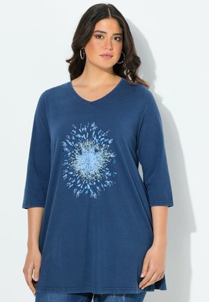 Vrouw in een marineblauwe tuniek met een lichtblauwe bloemenpatroon, driekwart mouwen en ringoorbellen, staand tegen een witte achtergrond.