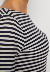 Zbliżenie na osobę w czarno-białą koszulę w paski, ukazujące dekolt i okolicę ramienia na jednolitym tle.