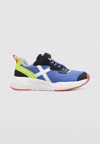 Zapatilla azul con acentos negros, logotipo blanco, detalles verdes y suela roja. Presenta una lengüeta de malla y una correa de Velcro para el cierre.