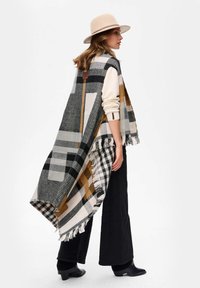 Długi, w kratę kamizelka w odcieniach szarości, bieli i złota z frędzlami, noszona na kremowym swetrze, w zestawieniu z czarnymi spodniami i szerokim kapeluszem.