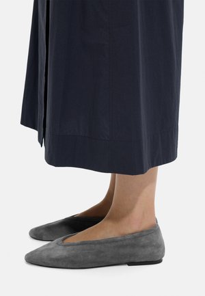 Szare zamszowe płaskie buty noszone z granatową spódnicą do kostek, pokazane z boku na jednolitym jasnym tle.
