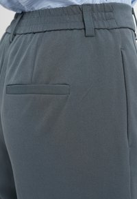 Pantalon en tissu gris avec une texture lisse, une ceinture élastique et une poche arrière unique au design plat.
