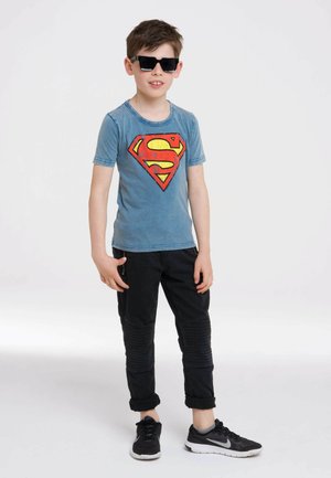 Blaues Baumwoll-T-Shirt mit einem großen roten und gelben Superman-Logo, kombiniert mit schwarzen Hosen mit gerippten Akzenten und schwarzen Sneakern.