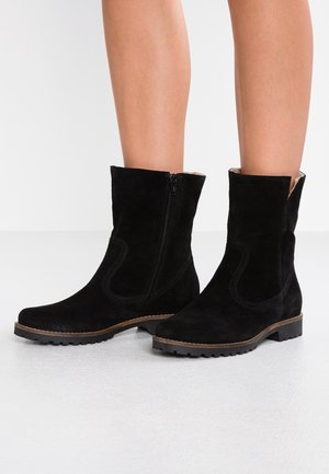 Czarne zamszowe kozaki do połowy łydki z bocznym zamkiem i brązową podeszwą, noszone przez osobę stojącą na jasnej powierzchni.