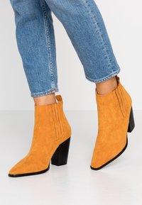 Bottines en daim orange avec un bout pointu et un talon noir épais. Caractéristiques : couture verticale sur les côtés et languette de tirage à l'arrière.