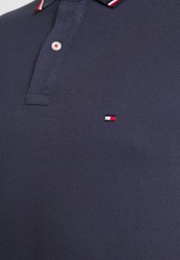 Polo bleu marine texturé avec un bouton blanc et un petit logo brodé rouge, blanc et bleu marine sur la poitrine.