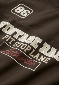 Camisa de algodón marrón con texto gráfico en blanco y rojo, que presenta "86" y "LÍNEA DE PARADA DE CARRERAS VINTAGE AUTÉNTICA" en diferentes tamaños de fuente.