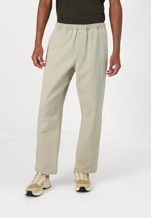 Mand iført beige bukser med elastisk talje og beige sneakers med hvide snørebånd stående mod en hvid baggrund.