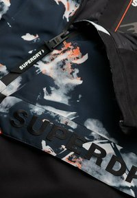 Veste noire en tissu texturé avec un motif abstrait orange et gris. "SUPERDRY" est embossé sur une étiquette noire près de la fermeture éclair.