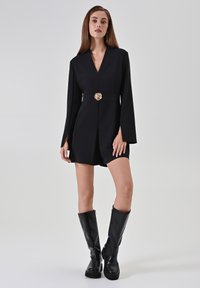 Una donna in piedi indossa un mini vestito nero a maniche lunghe con una cintura a fibbia a forma di fiore dorato e stivali neri fino al ginocchio su uno sfondo neutro.