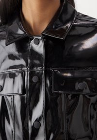 Black, glossy, faux leather jacket featuring a classic collar, button closure, two chest pockets with branded buttons, and stitch detailing.