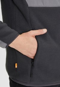 Fleecejas in donkergrijs met een contrasterend lichtgrijs bovenstuk. Voorzien van een zijritszak en een gestructureerde stof voor warmte.