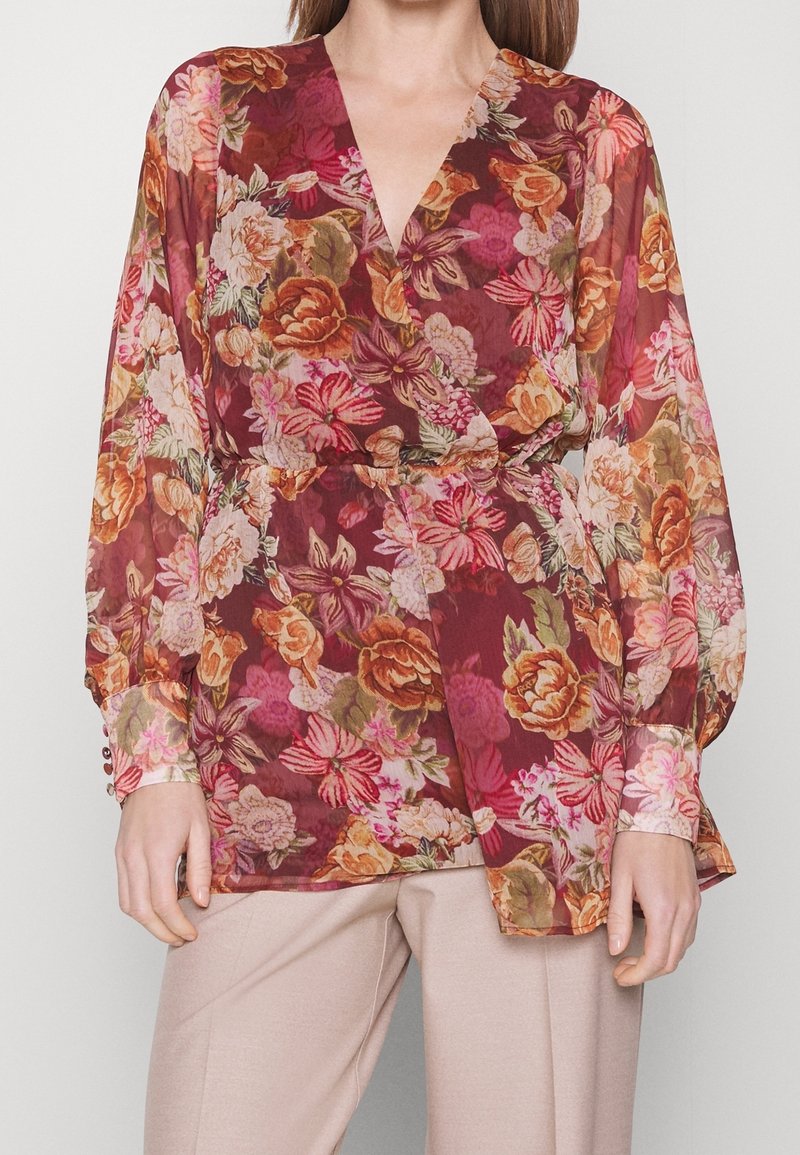 Femme portant un chemisier bordeaux à manches longues et col en V avec des motifs floraux roses, orange et verts, associé à un pantalon beige.