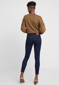 Sudadera marrón recortada con mangas anchas, combinada con jeans ajustados de color azul oscuro que presentan costuras icónicas en los bolsillos traseros y una etiqueta de cuero color marrón claro.
