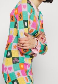 Un pull en tricot coloré avec un design en patchwork constitué de carrés roses, verts et jaunes. Agrémenté de diverses formes et textures.