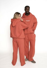 Două modele îmbrăcate în hanorace și pantaloni de culoare ruginie, unul întors cu fața spre cameră, iar celălalt arată spatele cu gluga ridicată.