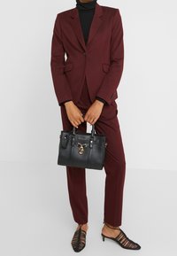 Černá kožená kabelka se zlatými detaily, obdélníkového tvaru, s výrazným uzamykacím prvkem, nesená v rukou v vínově červeném obleku na míru.