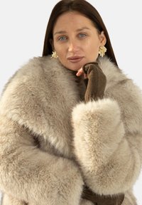 Beżowy płaszcz z sztucznego futra z szerokim kołnierzem o miękkiej teksturze. Modelka ma na sobie brązowe rękawiczki bez palców oraz kolczyki w kształcie złotych liści.