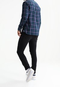 Camicia a quadri in verde scuro, blu navy e bianco, abbinata a jeans neri e sneakers bianche. Presenta maniche lunghe e polsini abbottonati.