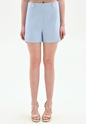 Femme portant un short taille haute bleu clair avec des détails à boutons et des sandales à talons hauts dorées à brides, debout devant un fond uni.