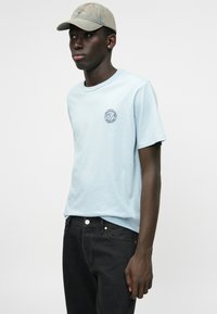 Camiseta de algodón de color azul claro con mangas cortas y cuello redondo, que presenta un pequeño logo oscuro en el lado izquierdo del pecho. Combinada con unos vaqueros oscuros.