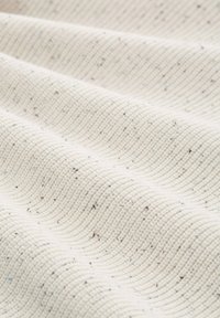 Strukturiertes Gewebe in einem cremefarbenen Farbton mit Wellenmuster, das kleine Sprenkel dunklerer Farben aufweist, die Kontrast und Tiefe erzeugen.
