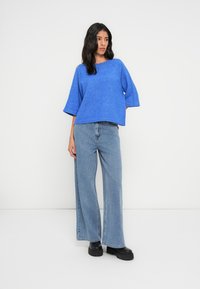 Pull court bleu à manches larges associé à un jean ample bleu clair. Le mannequin pose sur un fond uni, portant des chaussures compensées noires.