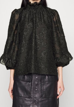 Femme portant un chemisier noir transparent avec des motifs floraux pailletés et des manches ballons, associé à une jupe en cuir noire boutonnée avec de grandes poches.