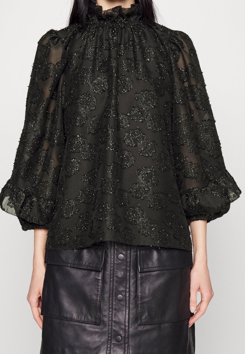 Femme portant un chemisier noir transparent avec des motifs floraux pailletés et des manches ballons, associé à une jupe en cuir noire boutonnée avec de grandes poches.