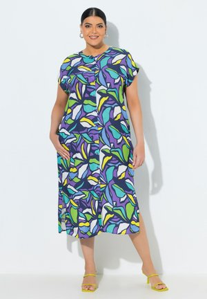 Donna taglia forte che indossa un vestito colorato a maniche lunghe fino al ginocchio con bottoni e motivi a foglie, abbinato a tacchi aperti gialli, in piedi contro un muro bianco.