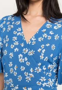 Robe portefeuille bleue en tissu texturé ornée d'un motif floral blanc et orange. Décolleté en V avec des manches courtes.