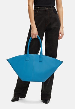 Blue leather tote bag with a unique angular shape, featuring two sturdy handles and a smooth texture. Black pants with a high waist.