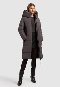 Duga puffer jakna u tamno sivoj boji s velikom kapuljačom, s patentnim zatvaračem, kviltanom teksturom i bočnim džepovima. Nošena preko crne suknje i tajica.