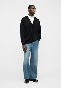 Άνδρας που στέκεται με τα χέρια στις τσέπες, φοράει μαύρη ζακέτα με κουμπιά, λευκό πουκάμισο, μπλε τζιν παντελόνι με φαρδιά πόδια και μαύρα παπούτσια σε απλό φόντο.