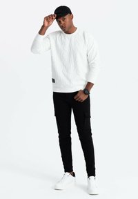 Vit textured sweatshirt med ett upphöjt mönster, matchad med svarta cargobyxor och vita sneakers. En svart keps bärs, med en klocka på handleden.