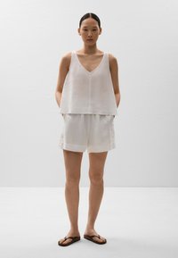 Top din in cu mânecă scurtă, alb din in, cu decolteu în V și croială lejeră, asortat cu pantaloni scurți. Sandale flip-flop maro completează ținuta.
