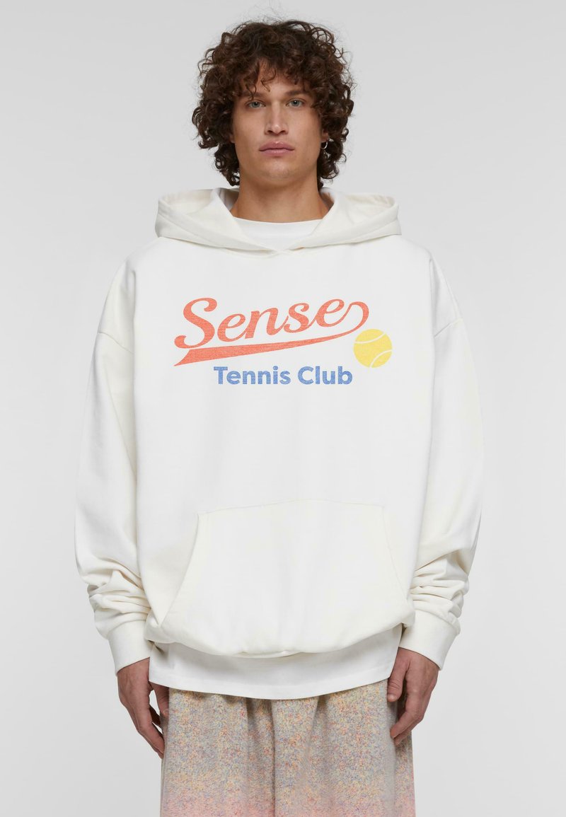 Balta kapuce ar priekšējo kabatu, uz kuras ir uzraksts "Sense Tennis Club" oranžā un zilā krāsā, ar tenisa bumbiņas grafiku, izgatavota no mīksta auduma.