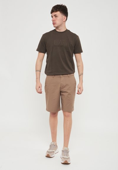 Maglietta marrone con logo in rilievo, pantaloni corti khaki e sneakers beige. Composizione in misto cotone, maniche corte e vestibilità rilassata con tasche laterali.