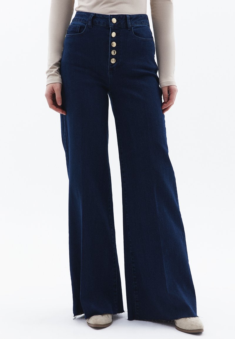 Džíny s vysokým pasem a rozšířenými nohavicemi v tmavě modrém denimu, s předním zapínáním na pět knoflíků a netkaným lemem na otvoru nohavic.
