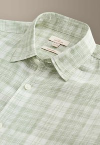 Chemise en lin à carreaux vert clair et blanc avec col boutonné et étiquettes visibles indiquant « Next Signature » et « 100 % lin ».