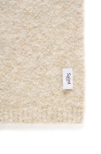 Tappeto in lana beige con una superficie testurizzata e un'etichetta piccola con la scritta "Saint" attaccata. Il design complessivo è semplice e minimal.