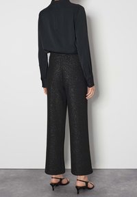 Kvinna i svart långärmad blus, glittriga vida byxor med hög midja och svarta öppna kittenhak-sandaler som står mot en vit vägg.