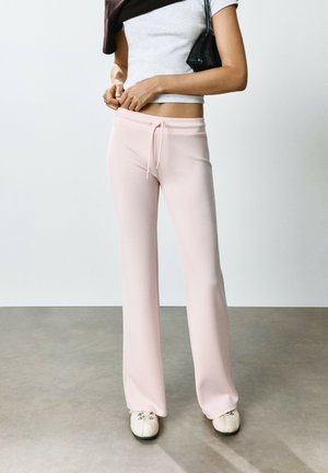 Femme portant un pantalon large rose clair, un haut blanc à manches courtes, tenant un sac noir sur l'épaule, debout sur un sol gris.